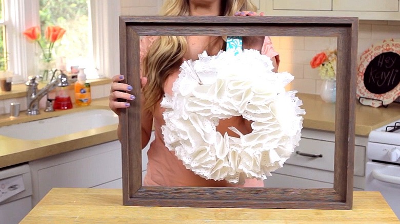 Creator displaying a wreath made of white paper doilies hanging inside of a wooden frame