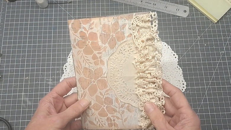 Crafter holding a journal with a paper doily on the front, a floral background, and a lacy edge