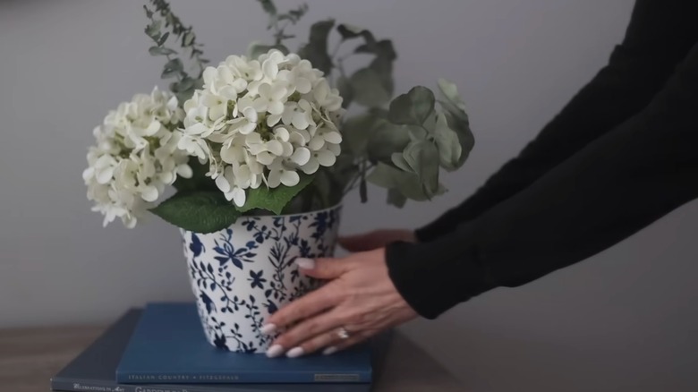 Person placing blue and white pot on a stack of books on a shelf