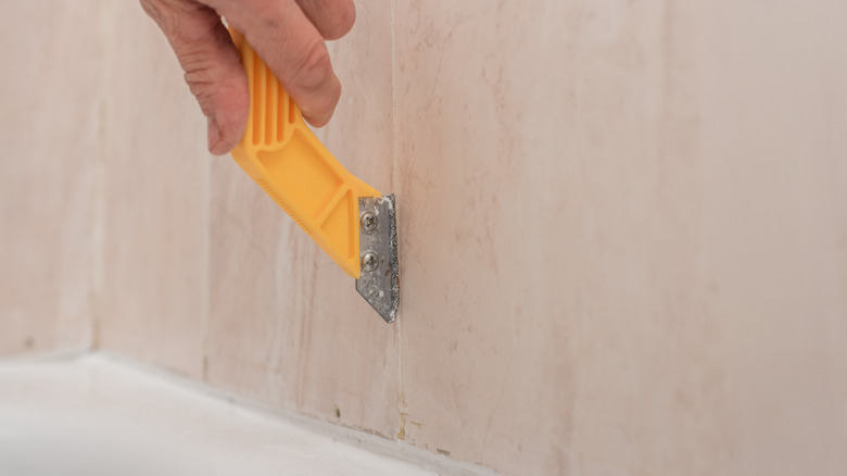A hand using a yellow caulk tool to remove silicone caulk from between tiles.