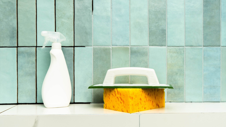 A sponge and spray bottle sit on a ledge in front of a tile wall with some of the grout lines resealed and some left unfinished.