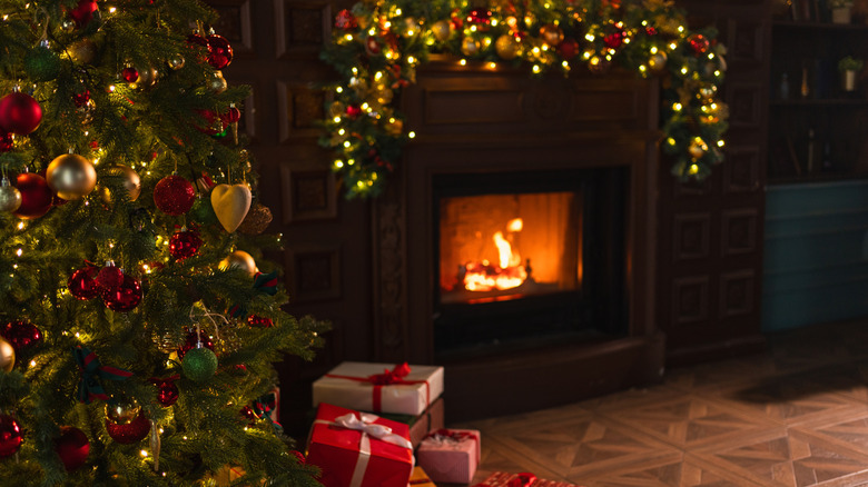 Side view of a Christmas tree with gold and red decorations along with gold glowing lights next to a fireplace with the mantle draped with a garland