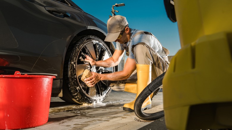 Man washing a black car's wheels with a red bucket nearby