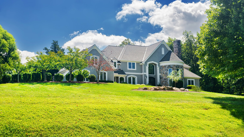 A sloping front yard in front of a large grey house with trees peppered through the property.