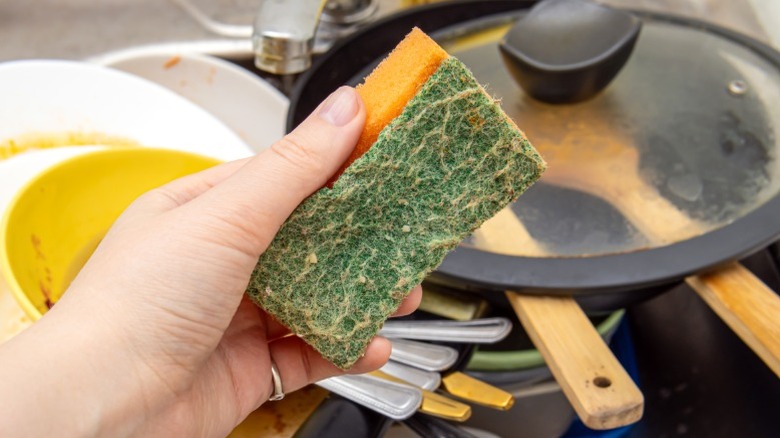 A person's hand holding old, worn-out sponge over sink of dirty dishes