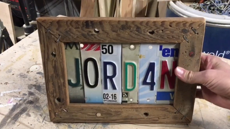 Hand holding a wood picture frame filled with slices of a license plate spelling out the name "JORDAN"