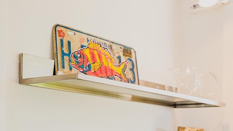 Weathered license plate with colorful fish painted on it perched on a shelf in a tiny house