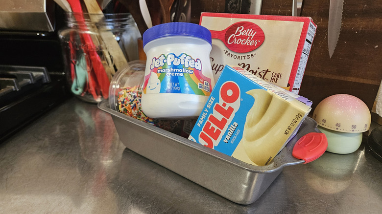 A loaf pan filled with baking ingredients including cake mix, pudding, sprinkles, and marshmallow cream