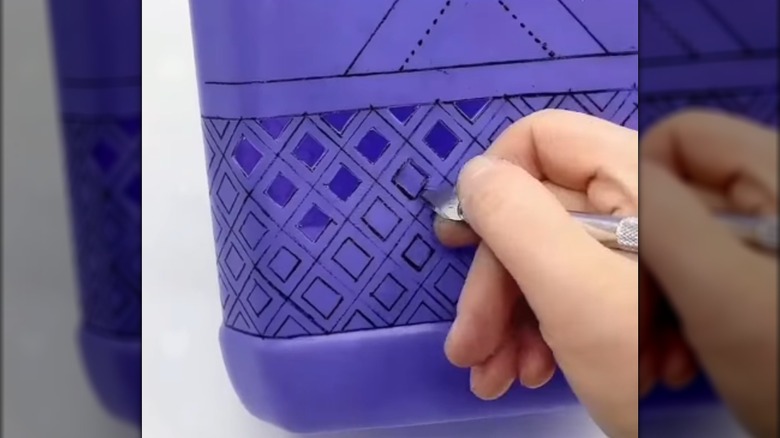 A light-skinned hand cuts a design into a plastic detergent container.