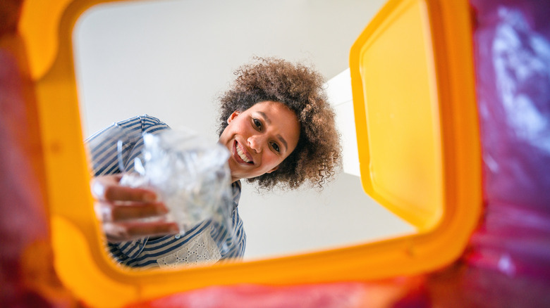 A smiling biracial woman places plastic in a recycling bin.