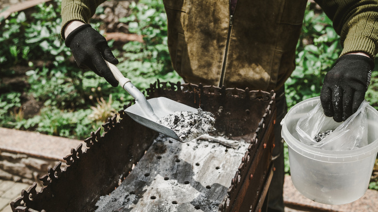 A person wearing black gloves scoops BBQ grill ash into a plastic bag inside a small bucket with a shovel.