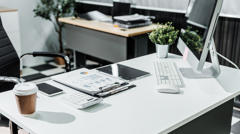 A modern white office desk with a plant, computer monitor, coffee cup, and work supplies