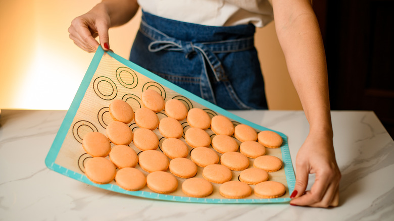 A woman holds a silicone baking mat with fresh macarons