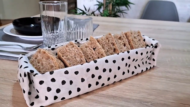 Black and white spotted tea towel made into bed basket holds bread on a table near drinkware