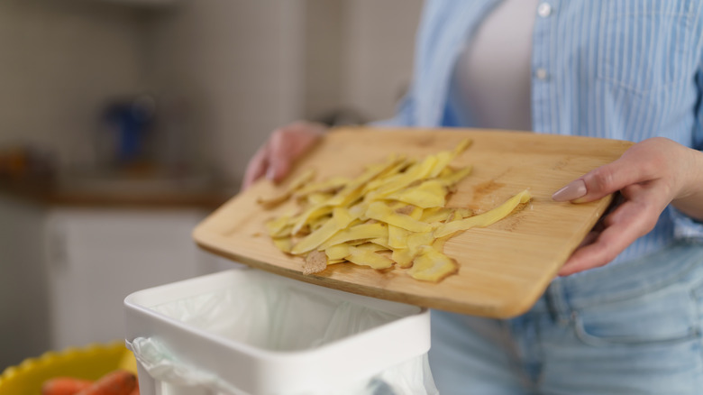 A person holding a wood chopping board with potato peels and tipping them into a small bin sitting on the counter
