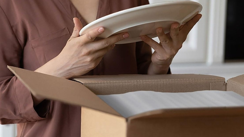 A woman holds a ceramic plate above a cardboard box filled with thin packaging foam sheets.