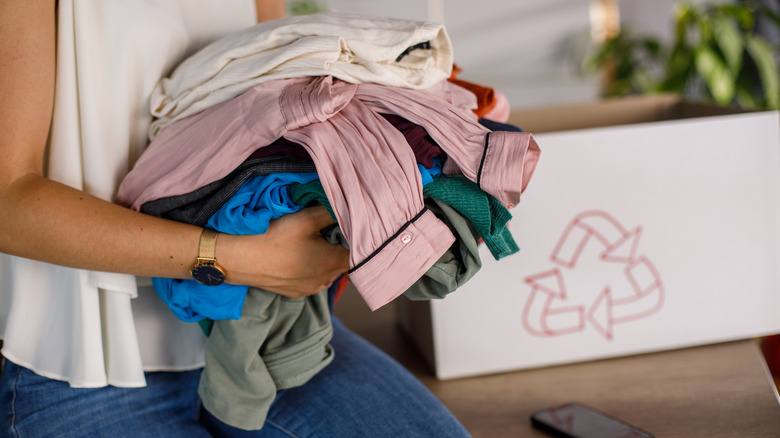 A person holding a pile of folded clothes with a box marked for recycling in the background