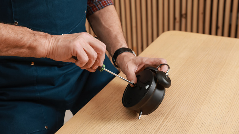 An older man repairs a black alarm clock using a screwdriver.