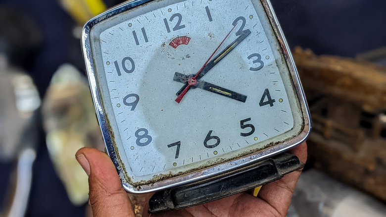 A person lifts  an old alarm clock out of a pile of trash.