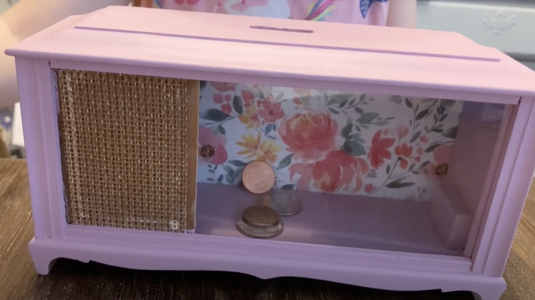 A money box made from a vintage wooden alarm clock painted pink and decorated with a wrapping paper background.