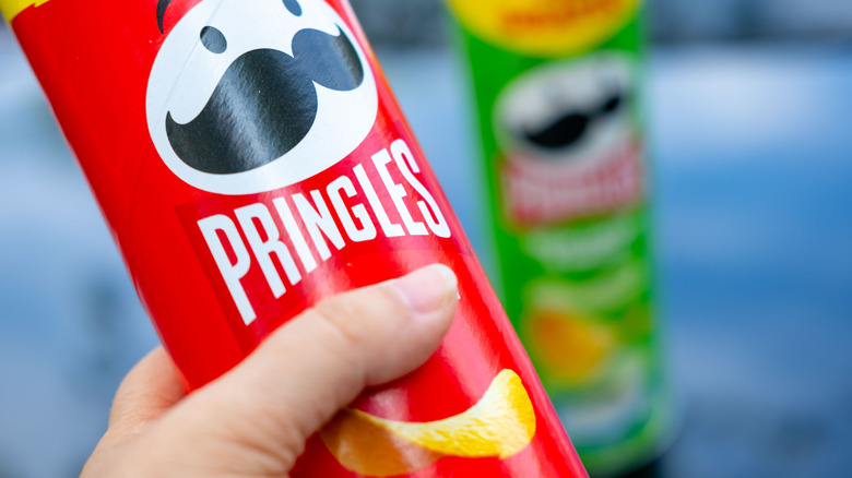 A hand holding a red Pringles container with a green Pringles tube blurred in the background.