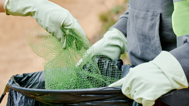 A person wearing gloves puts a green mesh produce bag into a black trash sack held open by another person wearing gloves.