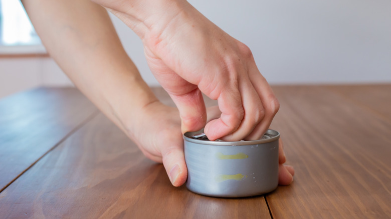A person uses the pull tab to open a tin can on a wooden tabletop.