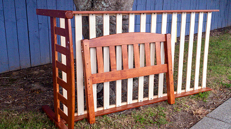 An old wooden bed frame leans against the trunk of a tree growing in a grass verge beside a sidewalk and in front of a blue fence.
