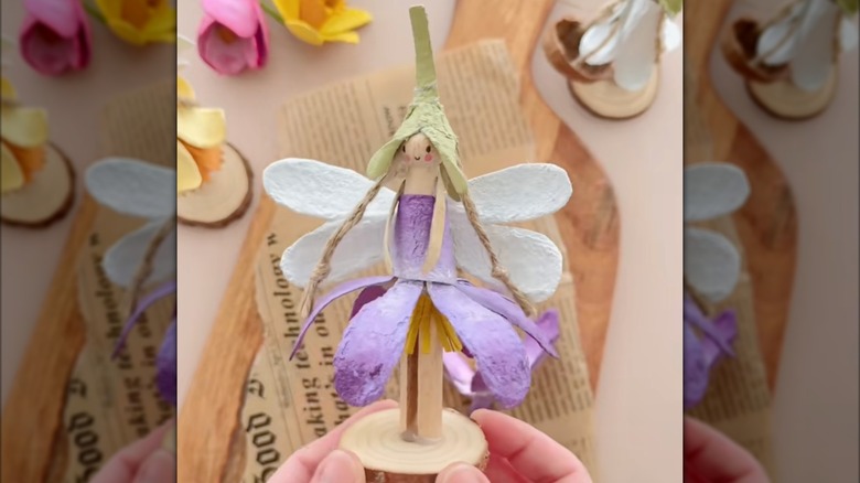 A wooden peg is decorated with white wings, a narrow pointed light green hat, purple dress, all made from an egg carton.