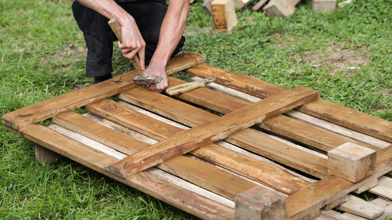 A person dismantles a pallet using a hammer and pliers in a grassy backyard.