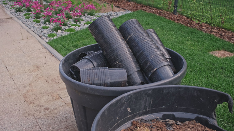 Stacks of black plastic nursery pots in a large container on a paved patio next to a lawn and a flower bed.