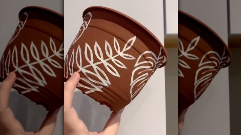 A hand holds a textured painted plastic flower pot with white leaf designs.