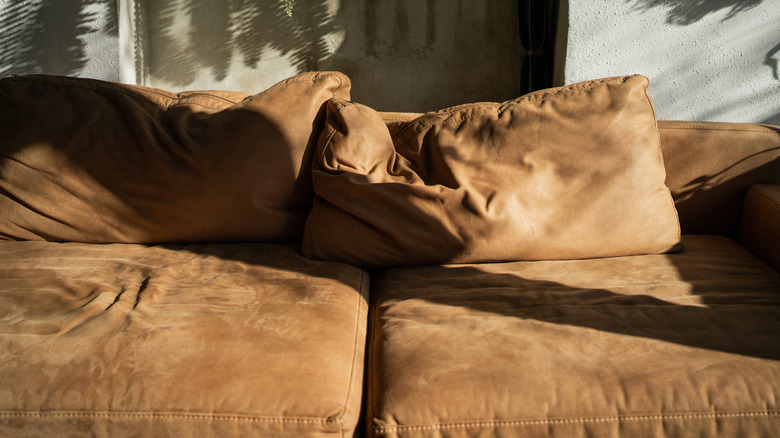 A brown leather sofa with worn-out cushions is dappled in sunlight on an outdoor patio.