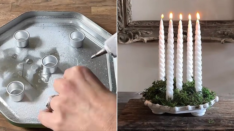 A hand glues a small metal cup to the inside of a cookie tin lid and a candle holder made from a cookie tin lid, moss, and white candles is set on a wooden credenza in front of an ornate frame.