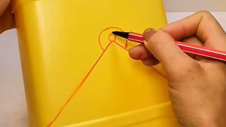 A hand draws lines in pen for cutting on a yellow plastic bottle.