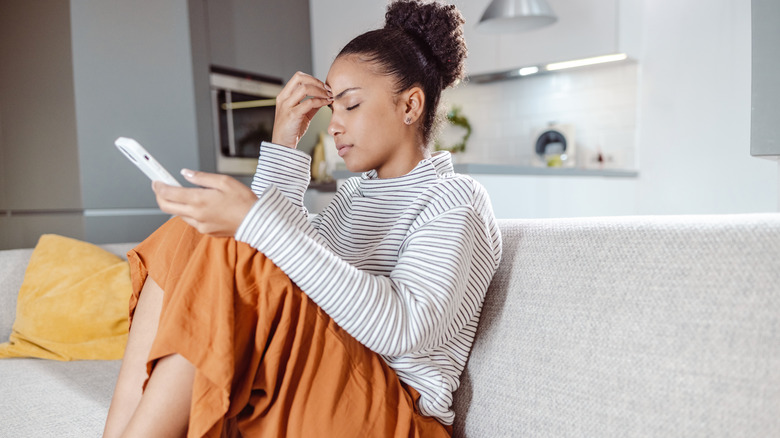 distressed young woman waiting on hold on smart phone on couch