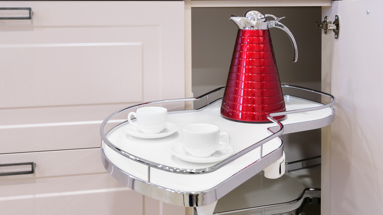 A small coffee pot and two white cups on little white trays on a pull out corner cabinet