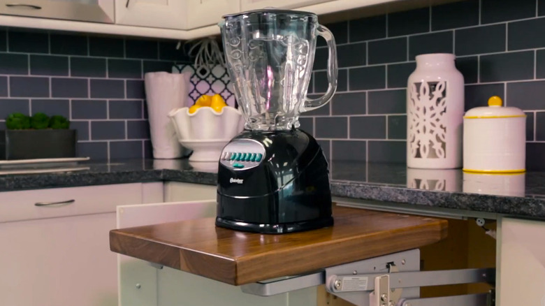 A blender on a wooden stand mixer shelf against a black counter and tile