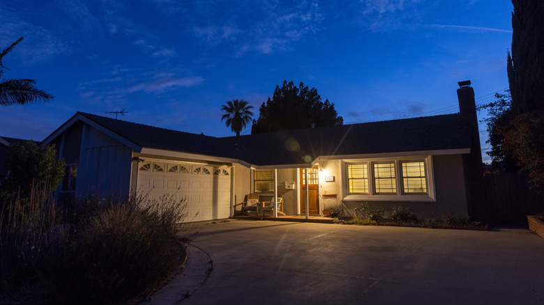 A white one-story L-shaped home is lit by a light next to the front door, while the driveway and front yard are in shadows. A dark blue sky and silhouettes of trees indicate that the sun has gone down.
