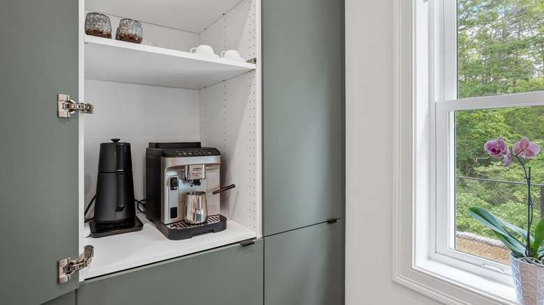 An open built-in cabinet displaying an espresso maker, mugs, and kettle on the shelves.