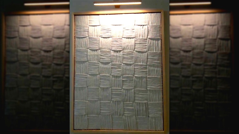 A handmade woven yarn wall art piece is framed and illuminated.