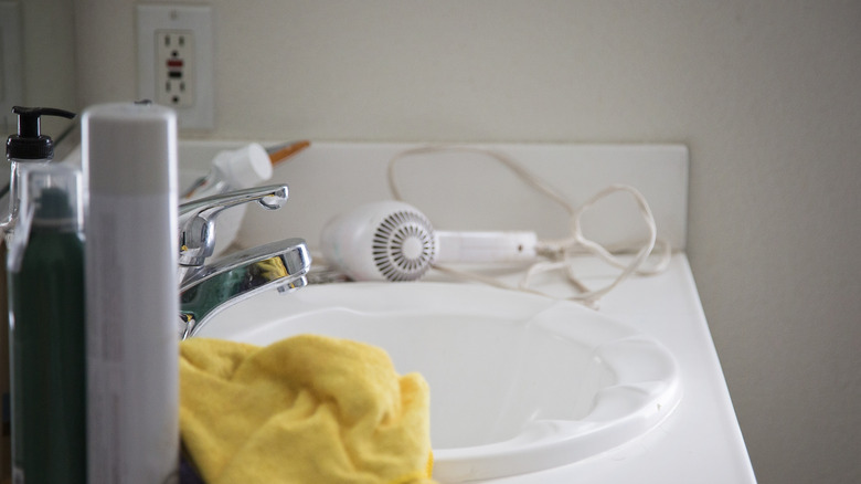 Cluttered bathroom countertop with a hairdryer, products, and a face towel