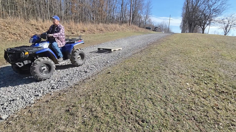 Man in a blue hat and flannel shirt riding a blue ATV dragging a wood pallet over a gravel driveway.