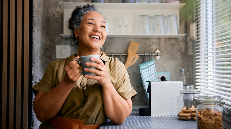 A homeowner enjoying a cup of coffee in a homey kitchen looking out the window, open shelving behind.