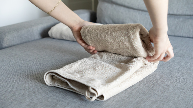 A person folds a beige bath towel on a gray couch