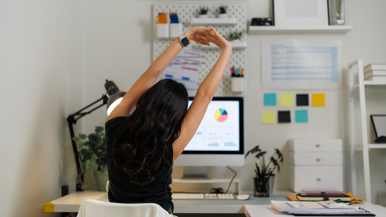A person stretches with their arms over their head while working at a computer in an at-home office