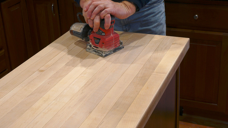 A person uses a sander on an island's wooden countertop to refinish it