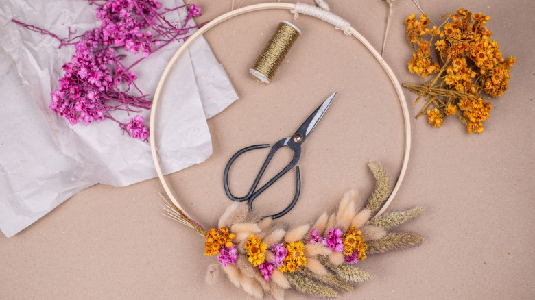 A wreath made of dried flowers next to gold thread, scissors, and piles of flowers