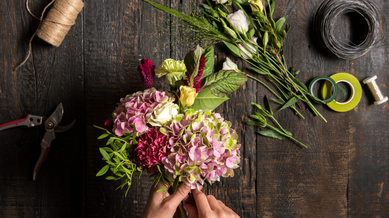 Top-down view of floral supplies on a wooden table. Two hands put together a floral arrangement.