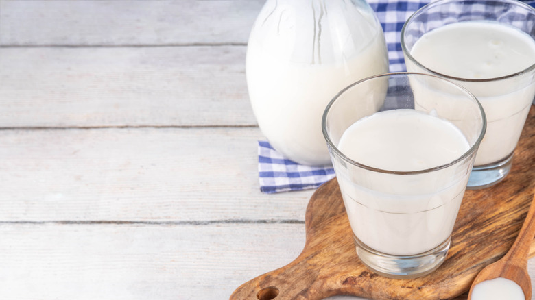 Glass of buttermilk placed on a wooden board with a ceramic jug and a blue checkered cloth in the background